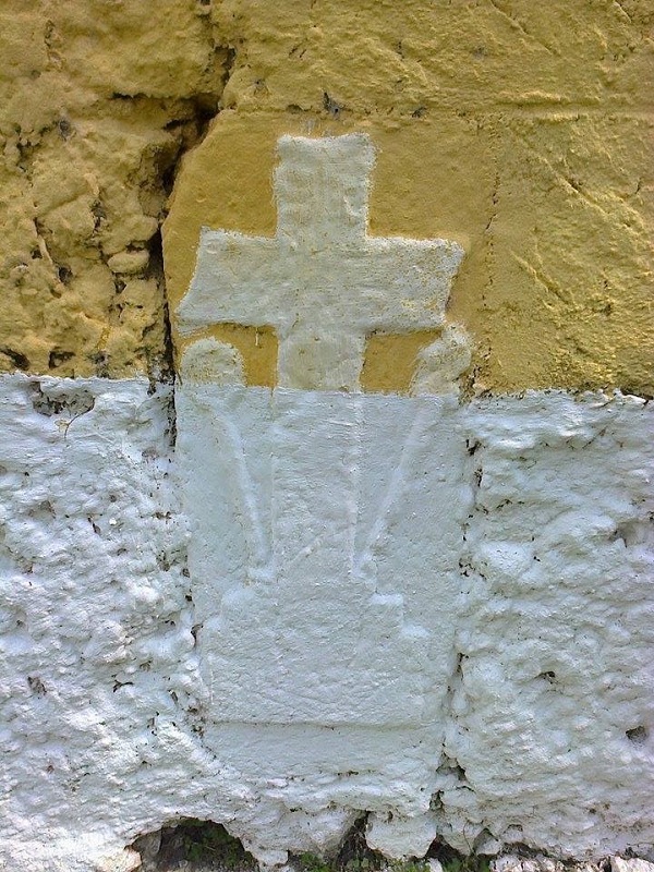 Detalle del monumento funerario adosado a la pared. Se aprecian dos símbolos; la banderilla y el estoque, dice la tradición local y sobre el relieve de la cruz, se aprecian otros detalles, irreconocibles hoy por años de pintura de cal sobre la superficie. Obsérvese con detenimiento la base de la cruz, los contornos de un cráneo, el trabajo de rescate cuidadoso, de estudio e identificación de los símbolos, nos dará o servirá para reinterpretar correctamente y objetivamente, la simbología de los decorados, quizá de la época colonial. (Foto Juan José Caamal Canul)